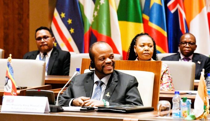 His Majesty King Mswati III smiles during the OACPS resource mobilisation session as Minister of Foreign Affairs Pholile Shakantu looks on from behind.